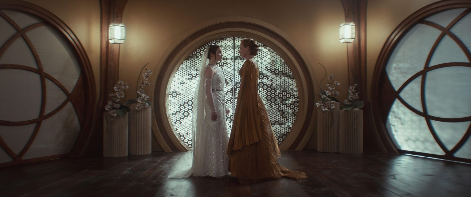 A woman in a brownish-orange dress stands facing a young girl in a white bridal dress in the center of an ornate brown room.