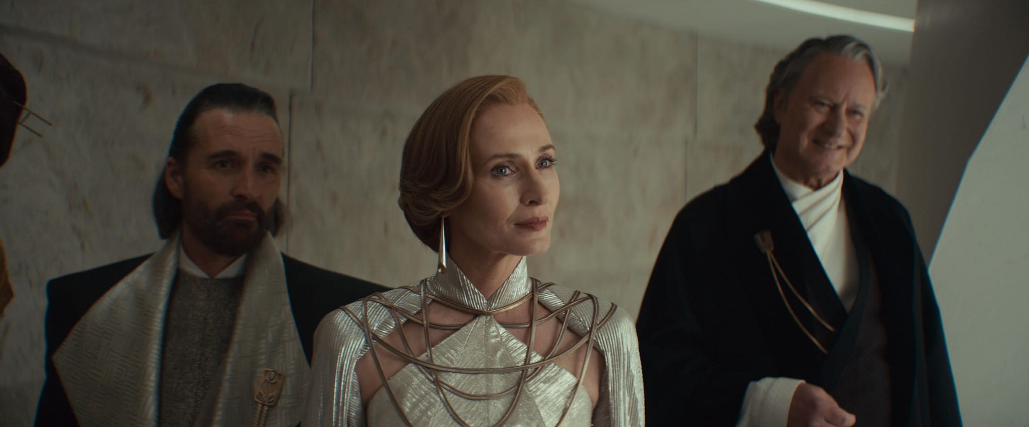 A woman in a ornate white dress stands in a room flanked by two men in formal robes looking at someone off screen.