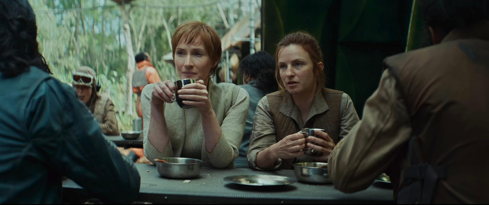 Two women, one in a white dress, the other in a brown and tan Rebel uniform, sit a table in a Rebel mess hall eating.