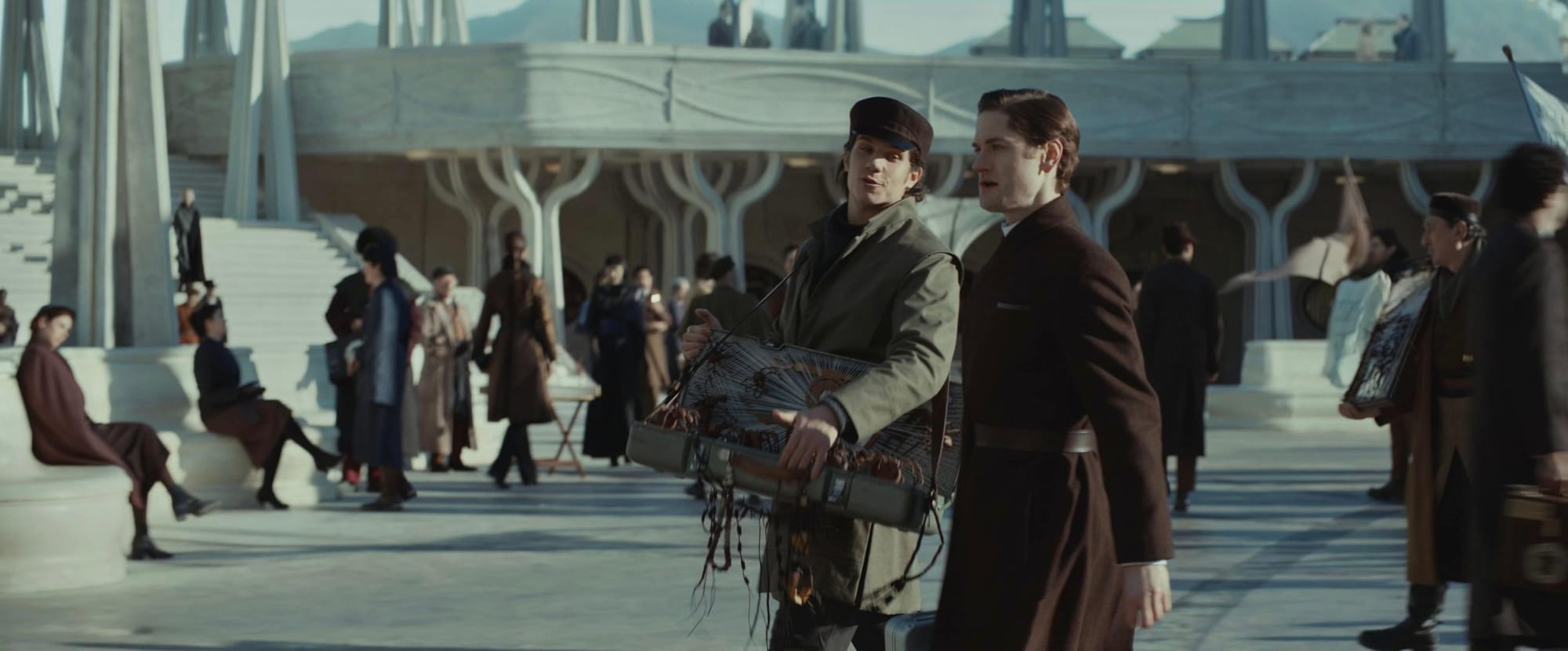 A man in a tan coat tries to pass a message to a man in a long brown coat in the middle of a city square