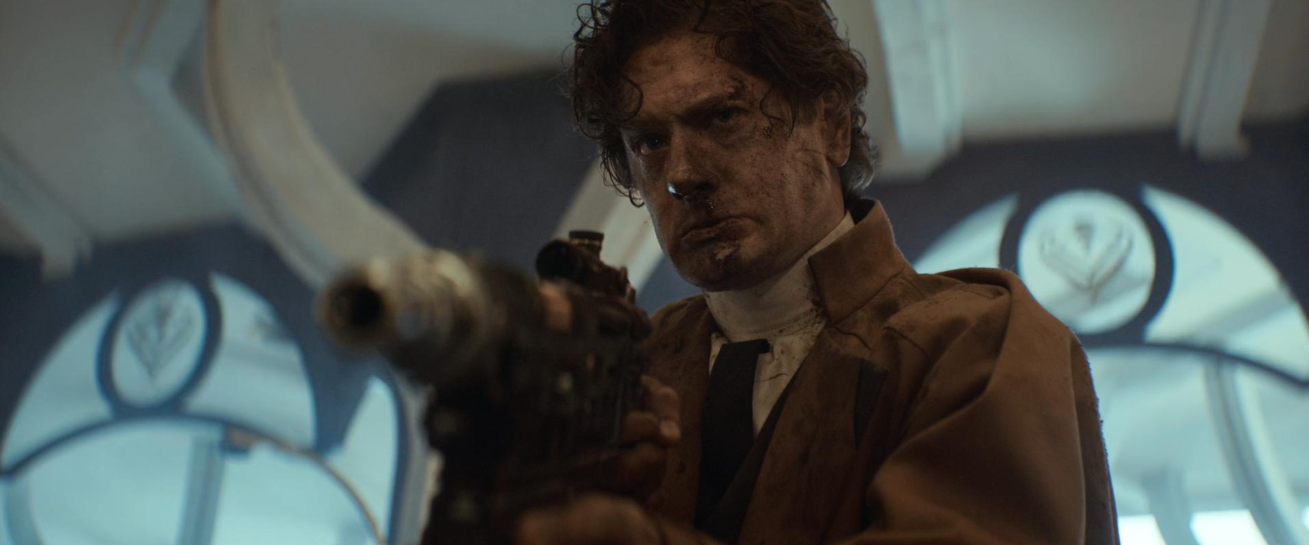 A man with a bloodied face holds a blaster while wearing a brown coat. He is taken aback by something he just heard.