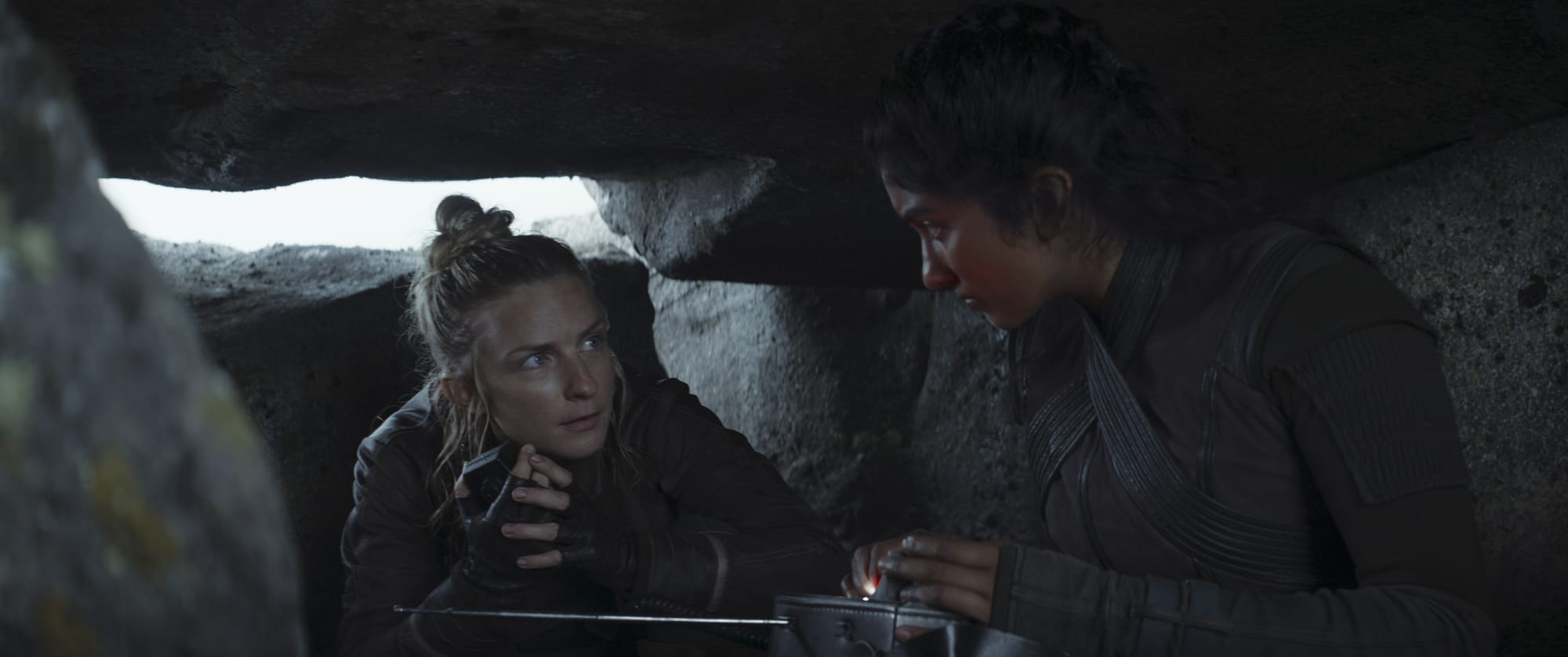 Two women in dark clothing huddle underneath rocks as they listen to a radio