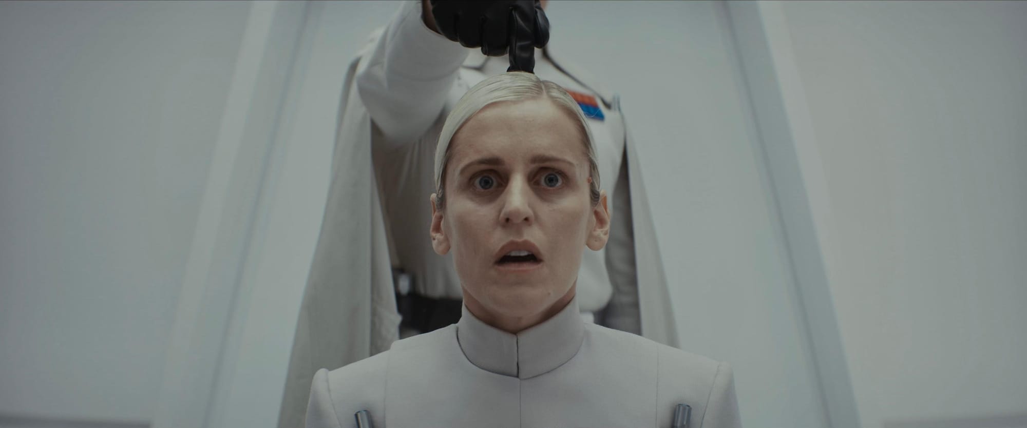 A woman with blonde hair sits while wearing a white coat. A man in a white uniform with a cape walks behind her and pokes her in the head.