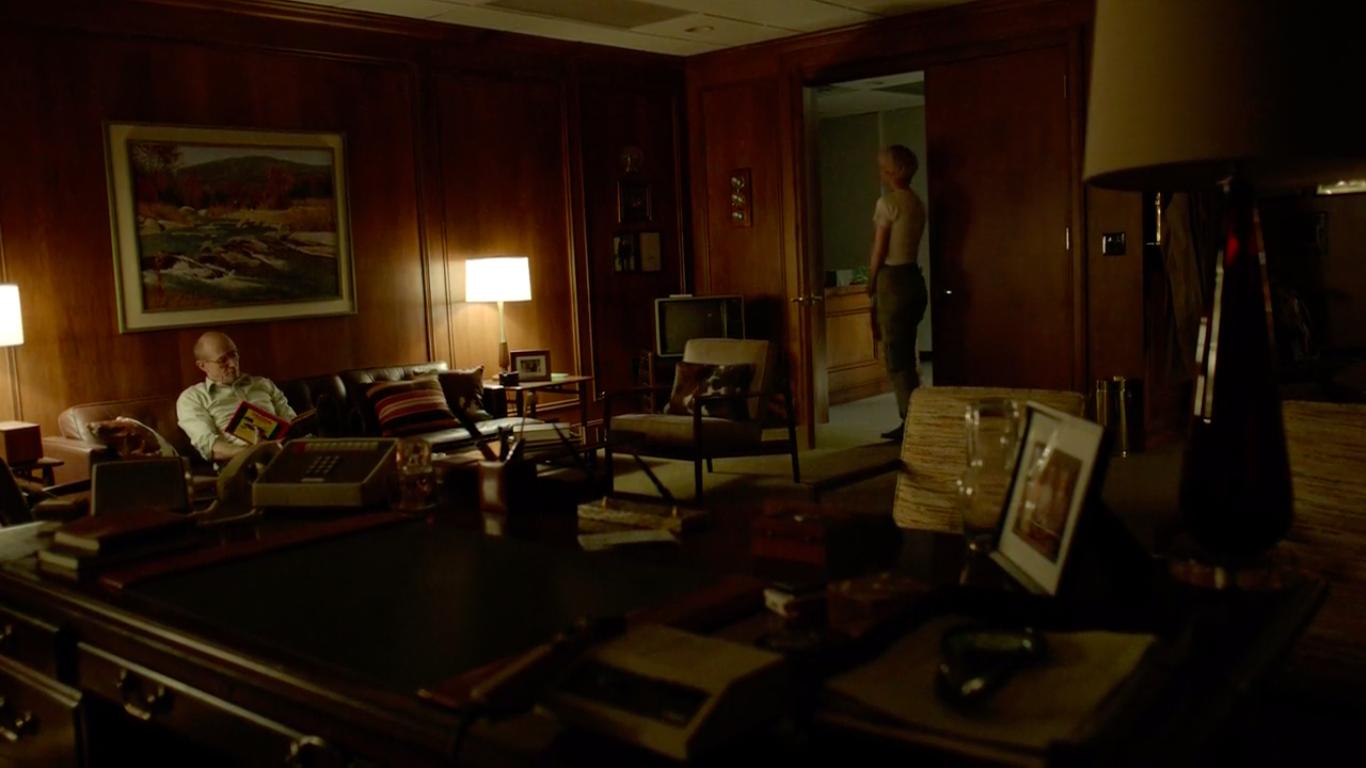 A middle aged man sits on a leather couch in his large office and talks to a young woman standing by the door.