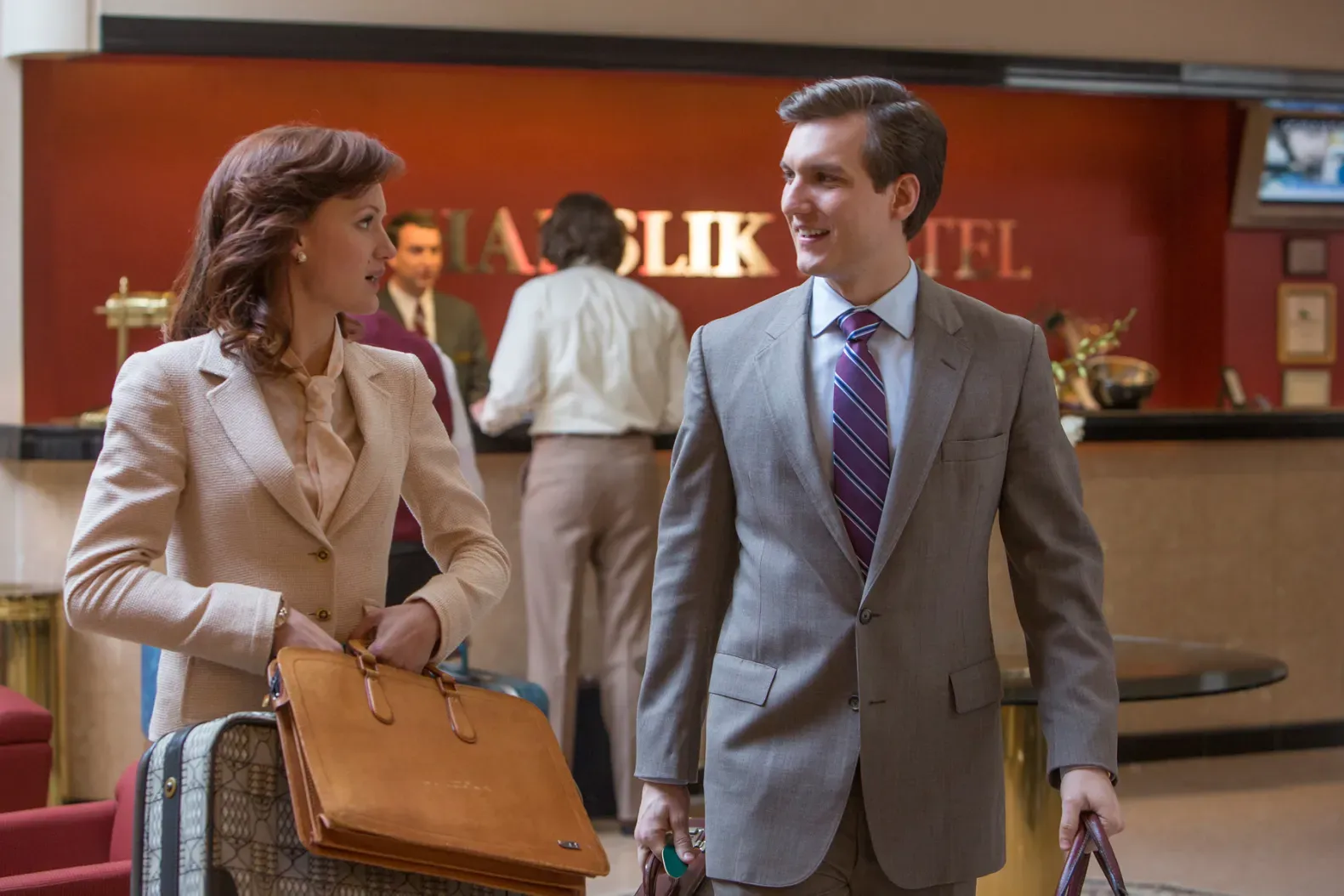 A woman in a tan suit and a man in a gray suit walk away from the front desk of a hotel with their luggage.