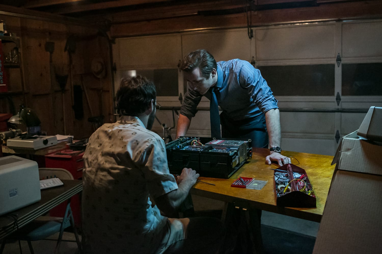 Two men look at a computer with its case open on a worktable in the middle of a garage.