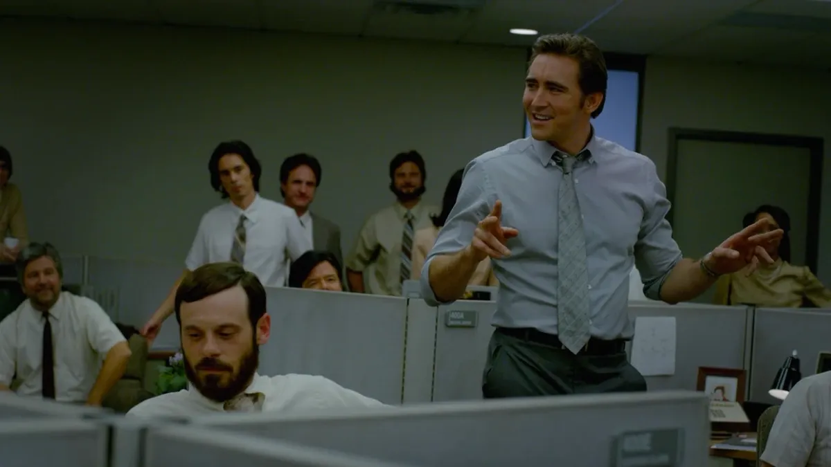 A man in a shirt and tie stands in the middle of an office floor and gives a speech to the assembled team.