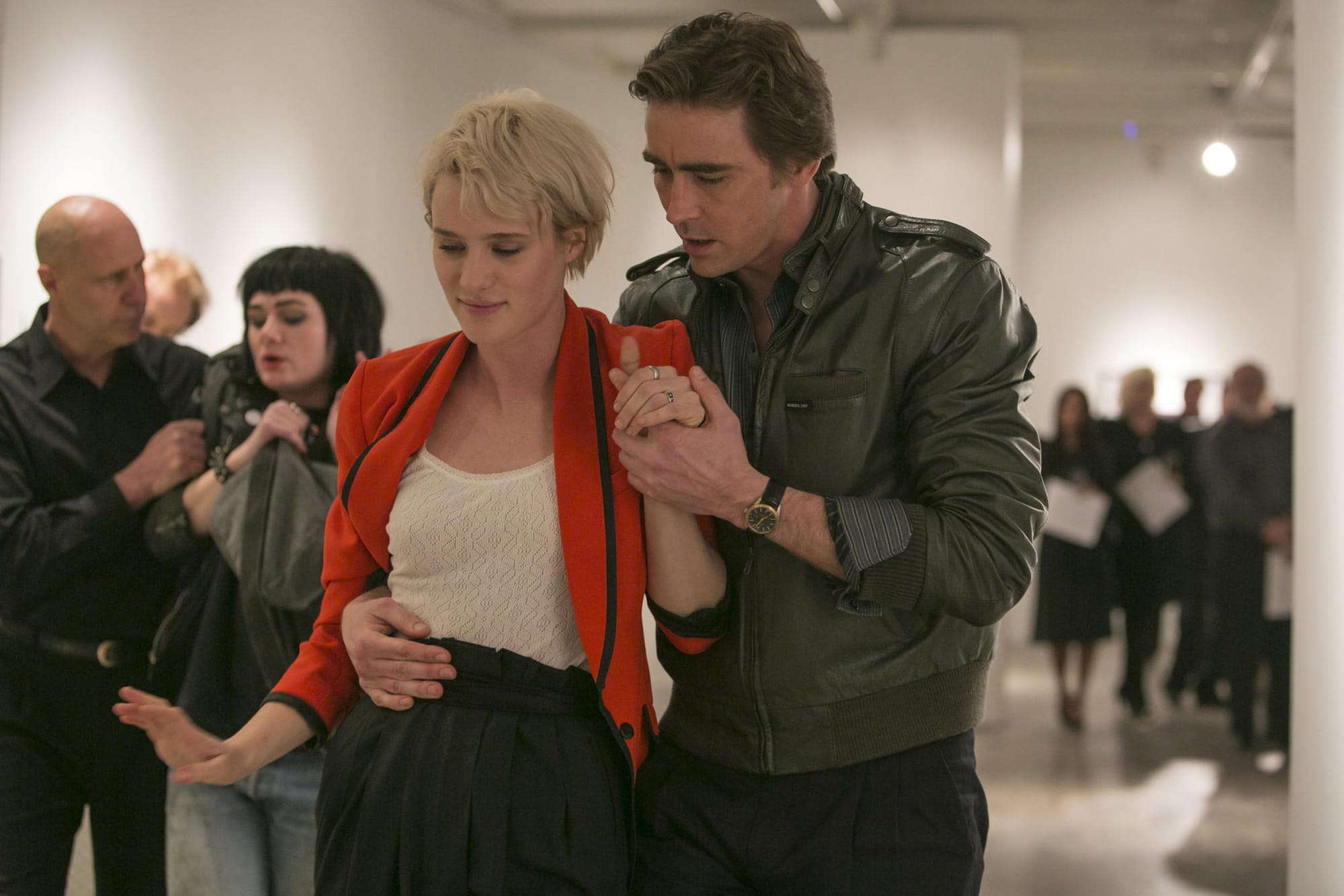 A man in a leather jacket helps a inebriated woman in a white top and red blazer leave an art show.