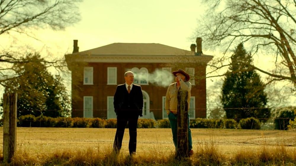 An old man in a cowboy hat smokes a cigar and talks to a middle-aged man in a dark suit. They are standing next to a barb wire fence.