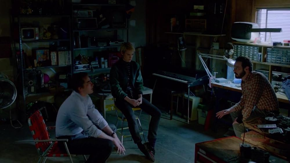 A man in a yellow and black flannel shirt sits in a dark garage. He is talked to a man in light shirt and woman in a dark sweater.