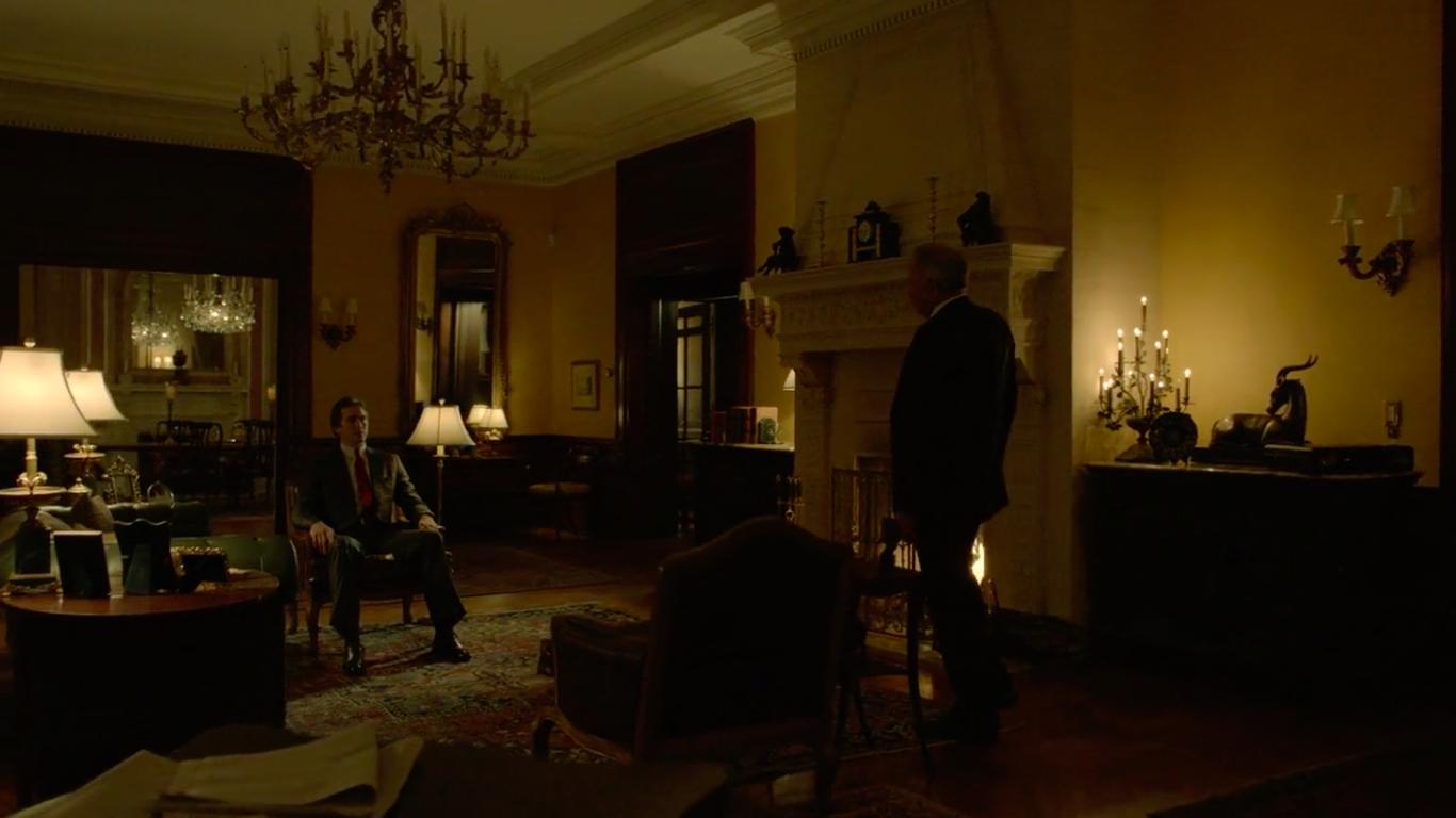 In an ornate and gigantic living room, an older man in a suit stands by a fireplace talking to a younger besuited man sitting down.