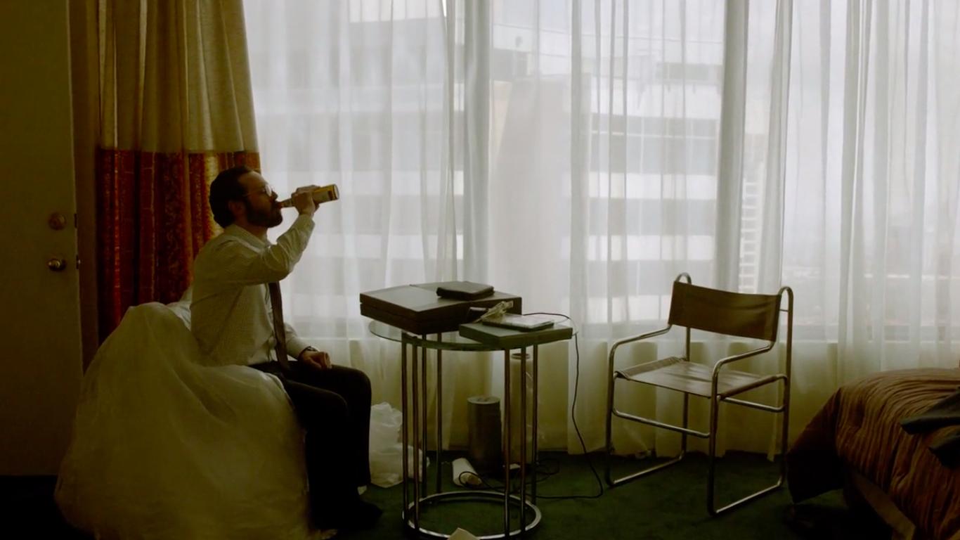 A man sits by a window in a hotel room. He is drinking a beer. A portable computer sits in front of him.