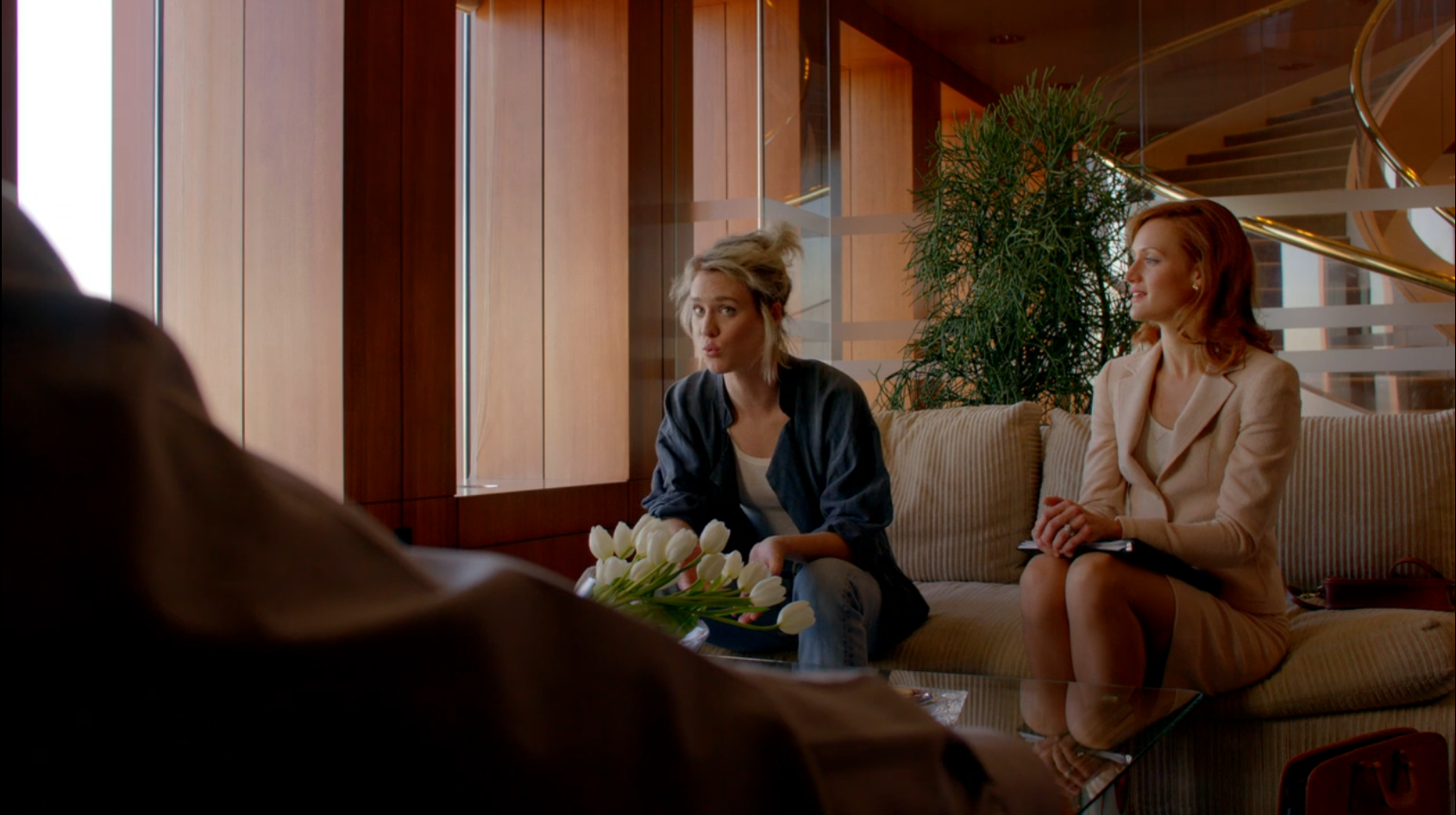 A woman in a tan suit sits next to another woman in ratty blue jeans and a wrinkled shirt as they pitch a venture capitalist.