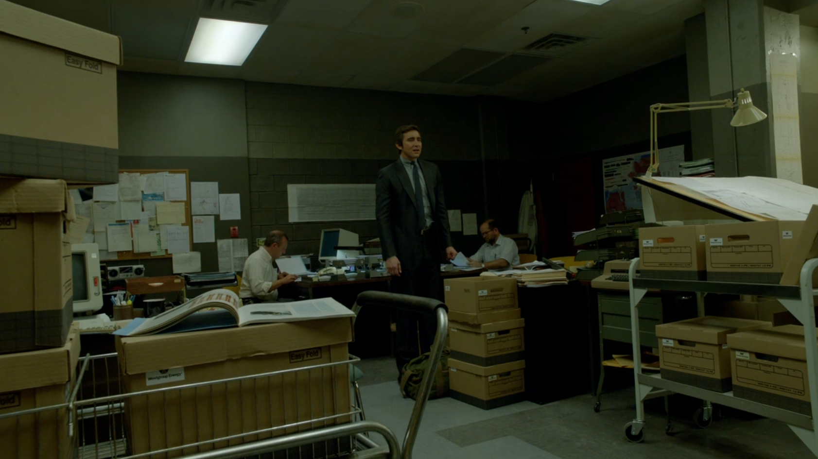  A man in a suit stands in the middle of a depressing office in a basement.