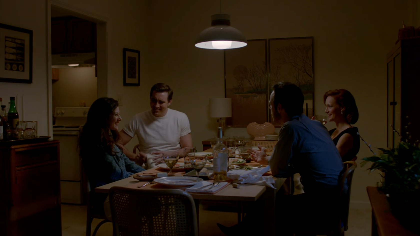 A group of four people eat dinner around a table. The mood feels casual yet guarded. The decor is straight out of 1985.