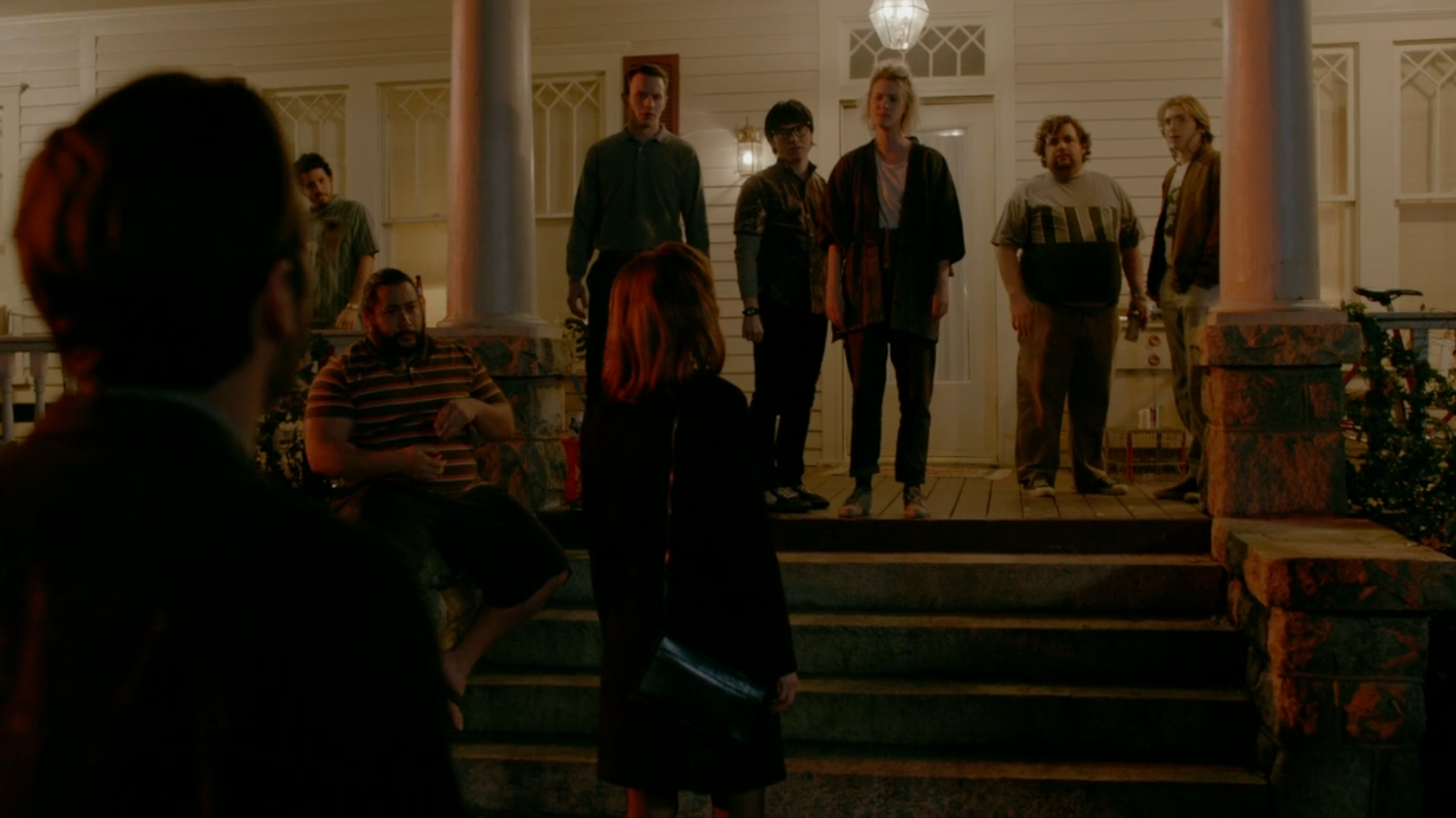Several people stand on a porch, looking warily at the woman in front of the house, and the man standing behind her.