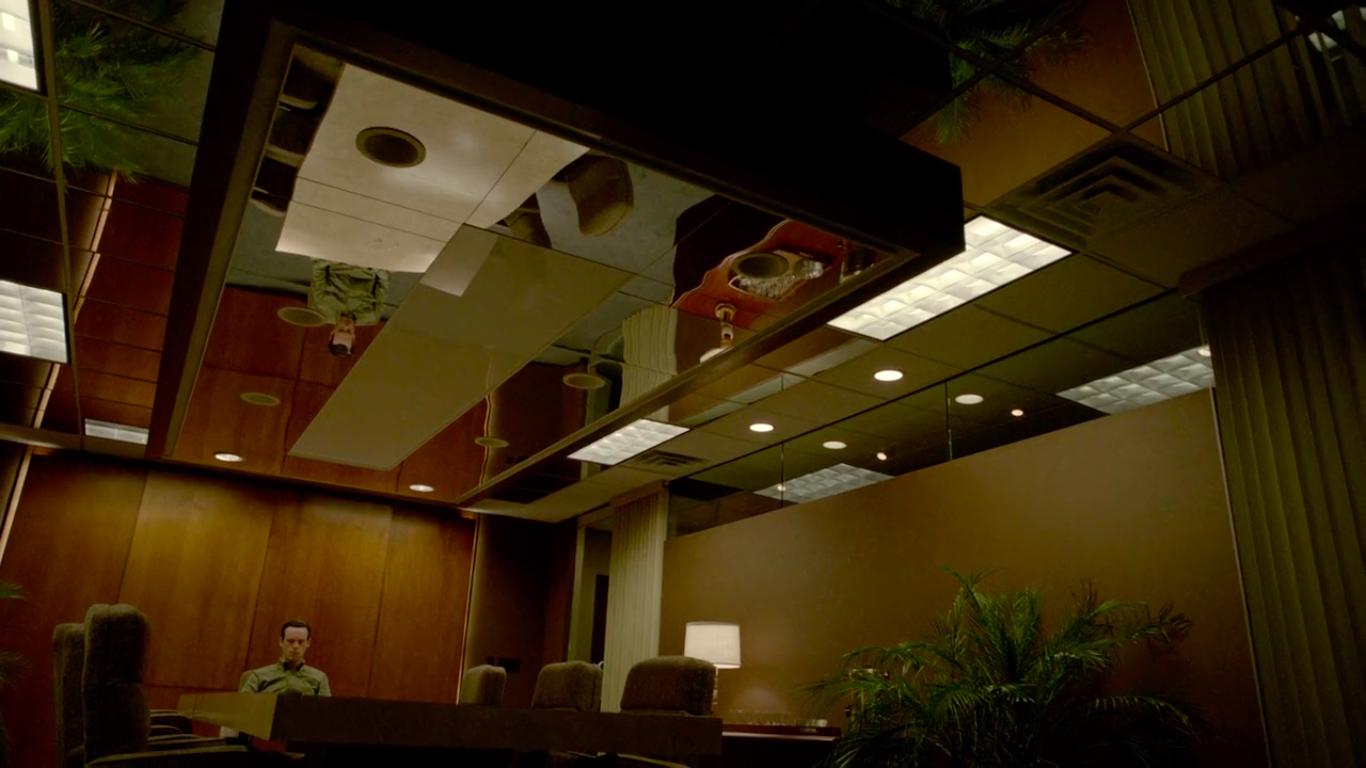 A man sits alone at the head of a conference table in a large conference room with a mirrored ceiling.