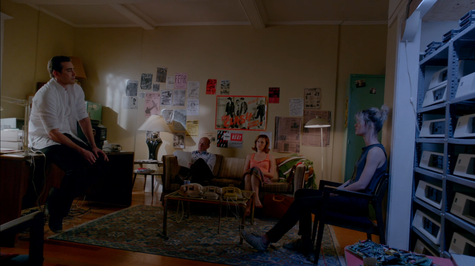 A man in a white shirt sits on a desk as he talks to a woman in chair. In the middle are a man and woman on a couch.