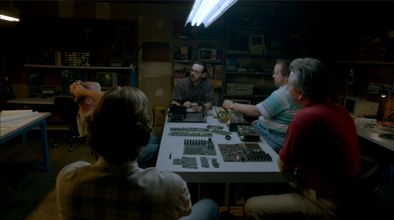 A man talks to a group of men around a work table. On the table are components for a computer.