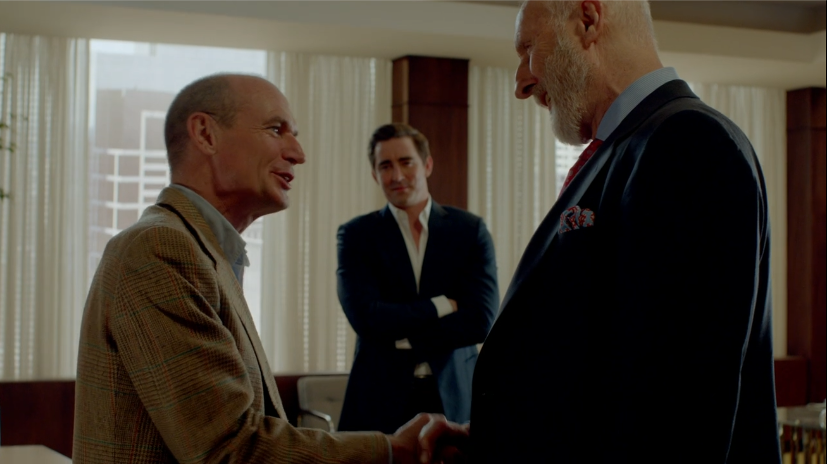 A man in a tan coat shakes hands with a taller man in a blue suit, as a third man stands in the background.