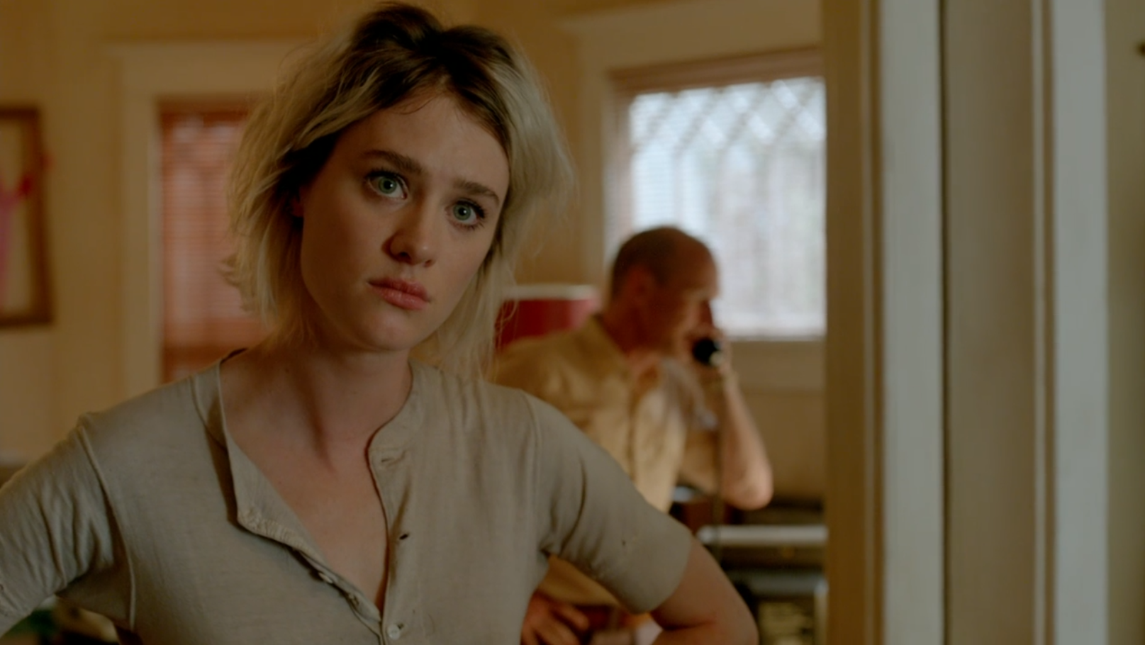 A young woman with dyed blonde hair holds her hands on her hips as she listens to people make choices as a man in the background answers a phone.