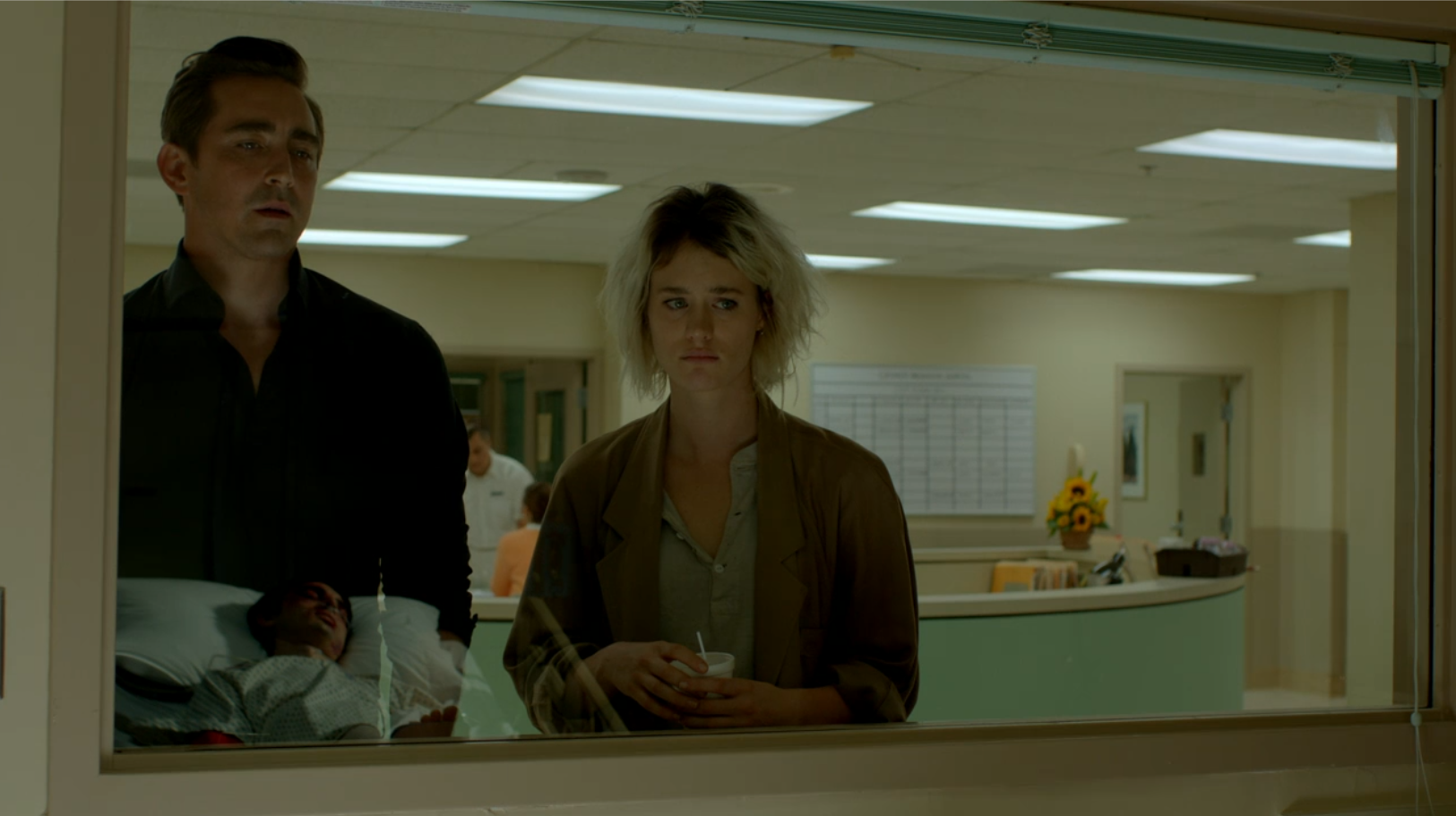 A man and a woman stand at hospital room window. In the window is a reflection of a man in hospital bed.