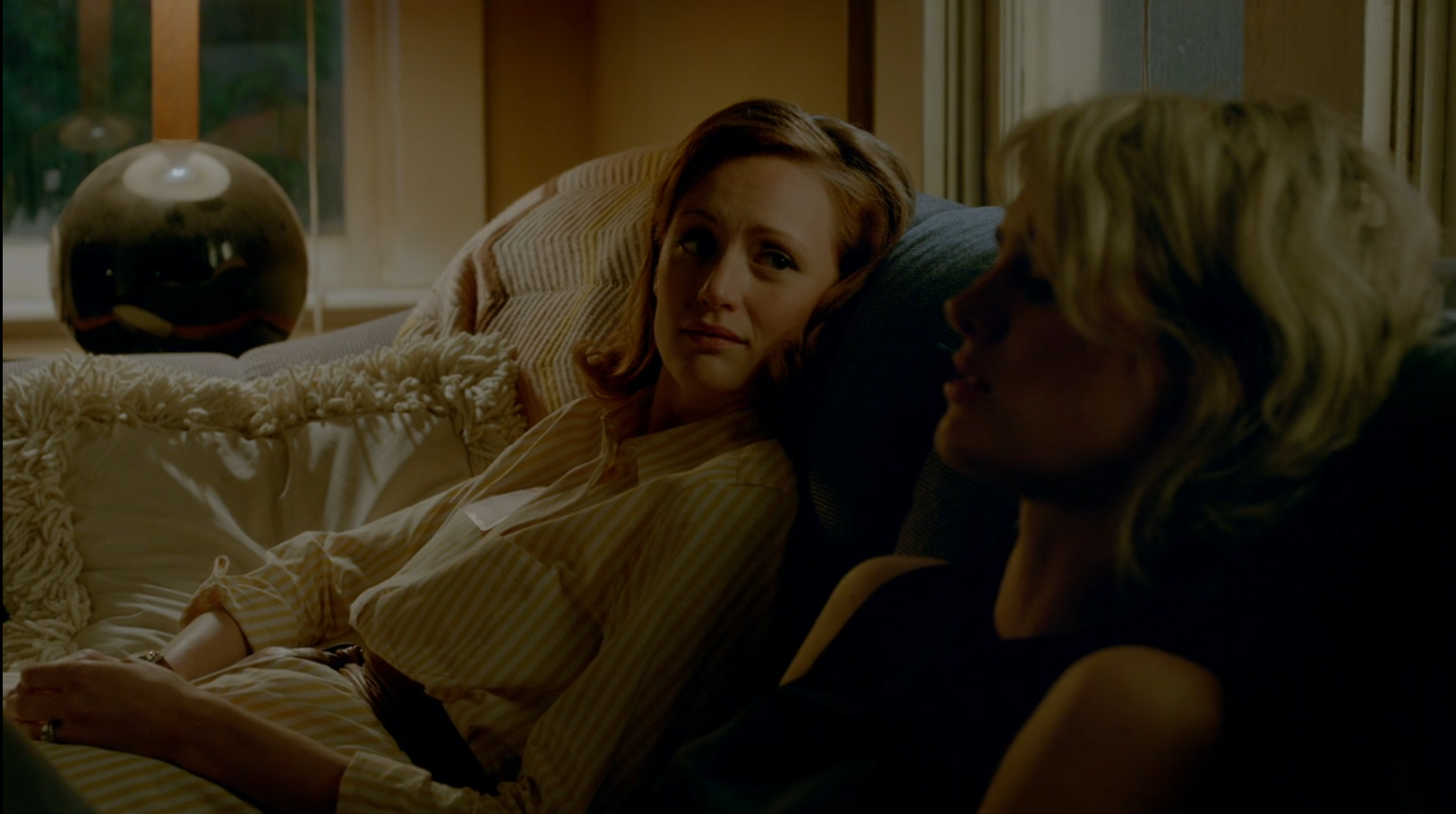 A woman with red hair and a woman with dyed blonde hair sit on a couch and talk.