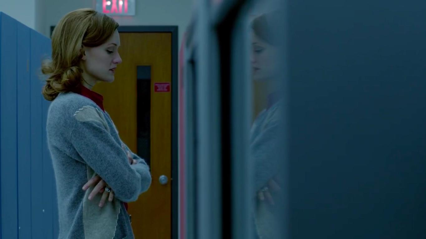 A woman with red hair wears a sweater and has her arms folded. She looks like she is thinking about something.
