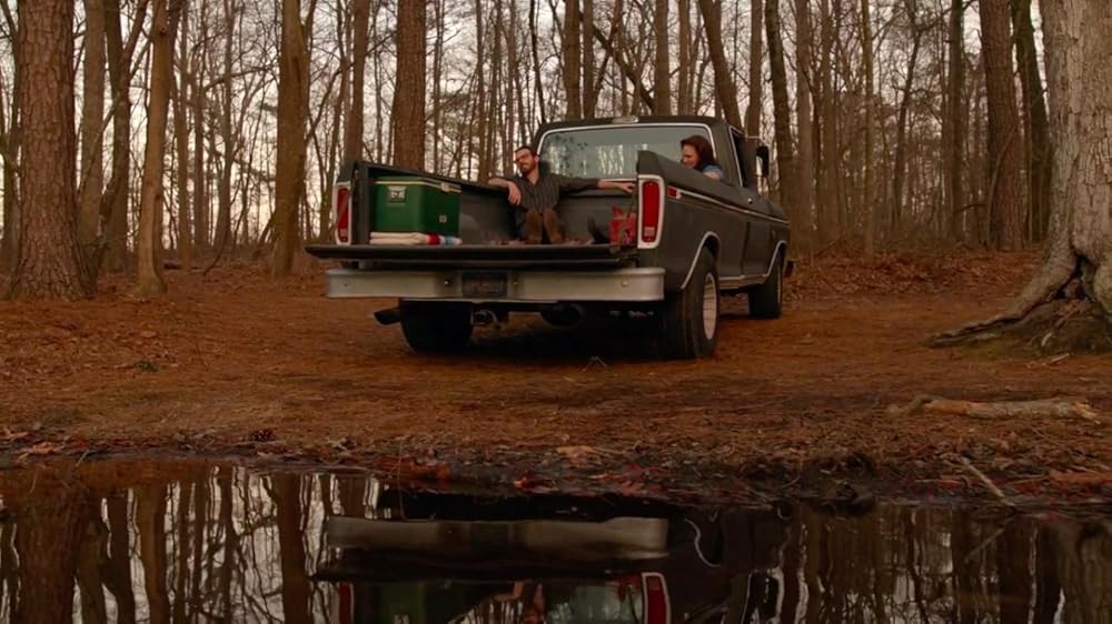 A man and a woman sit in the back of an old truck with a cooler and talk about things. The truck is parked by a lake.