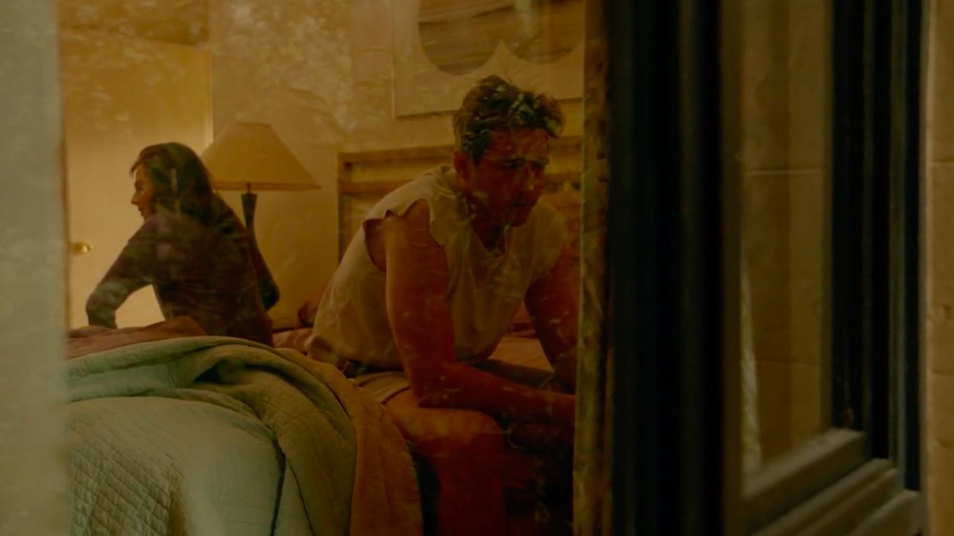 The camera is shooting through a window so we see a reflection. Inside, a man in a gray t-shirt and a woman in a pajama top talk, facing away from each other.