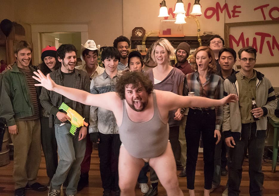 A picture several people reacting to bearded man in underwear and a gray tank top crashing the picture right as it clicked.