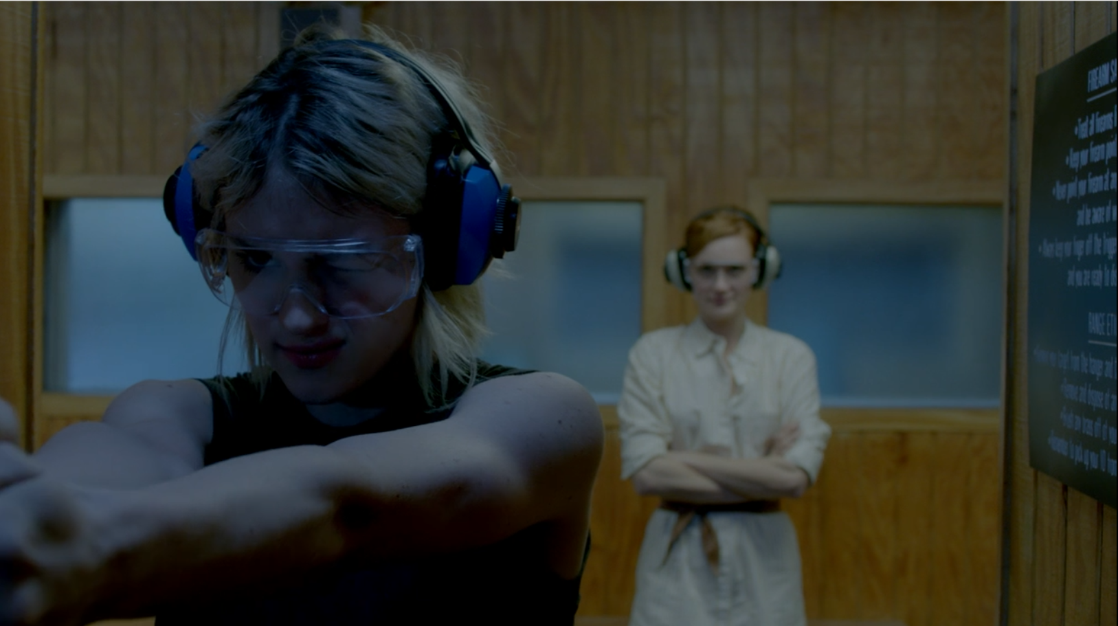 A woman with dyed blonde hair shoots a gun at an indoor shooting range as a woman with red hair watches in the background.