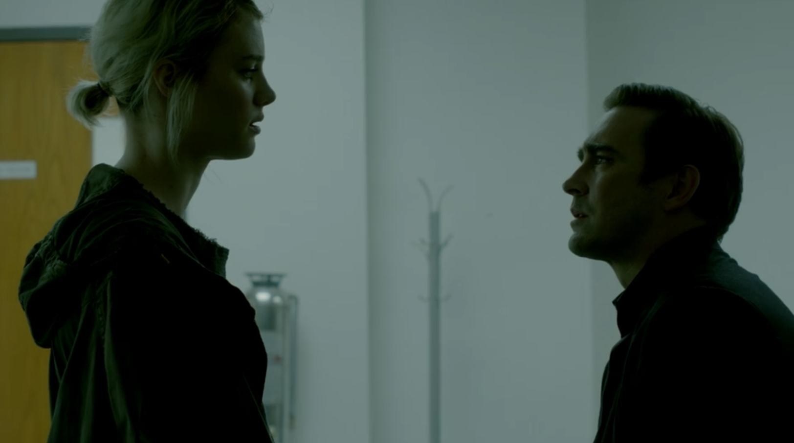 A young woman with dyed blonde hair looks longingly at a man with brown hair sitting on the edge of a desk.