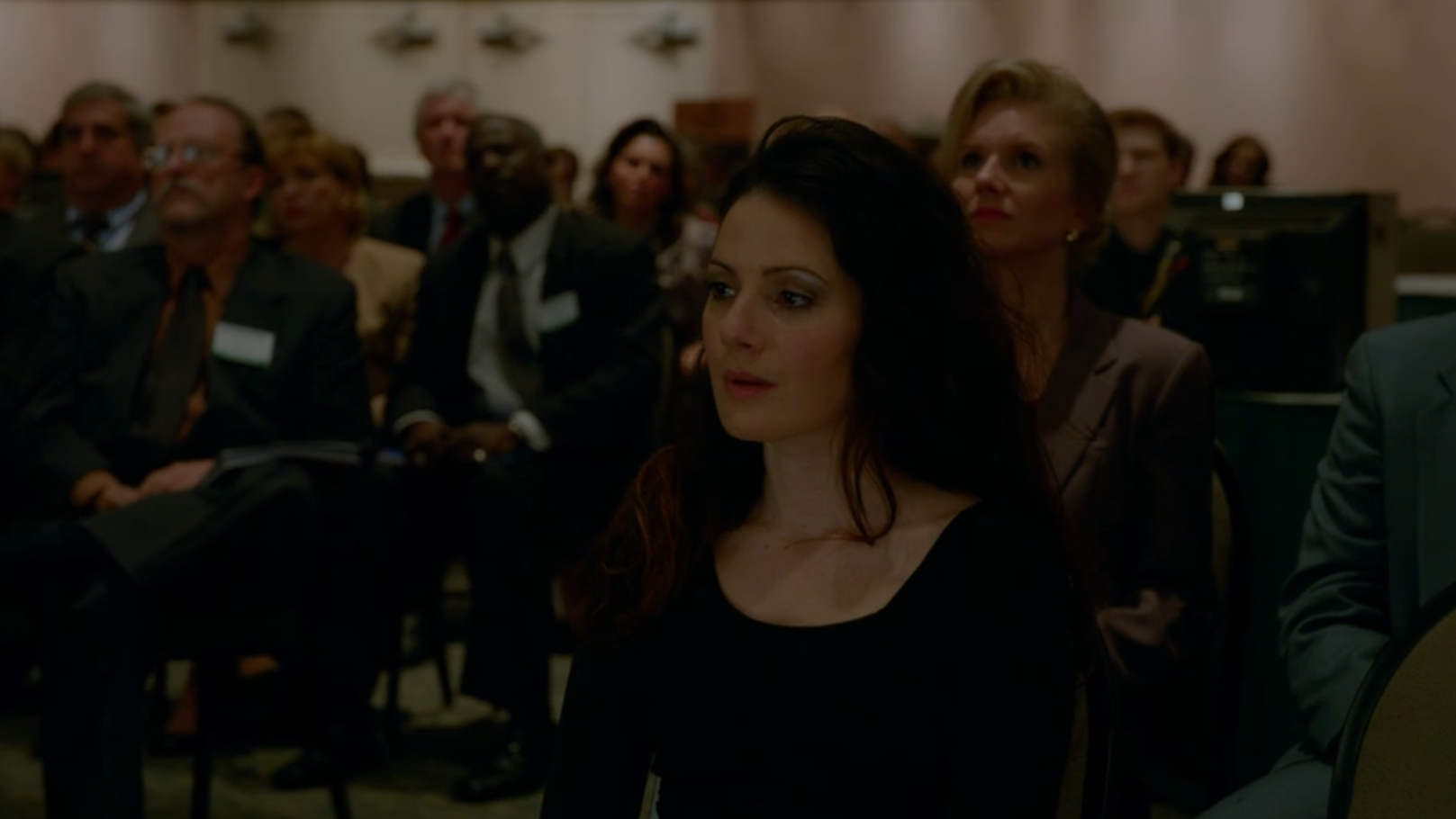 A woman with dark brown hair looks heartbroken as the people around her watch someone giving a presentation.