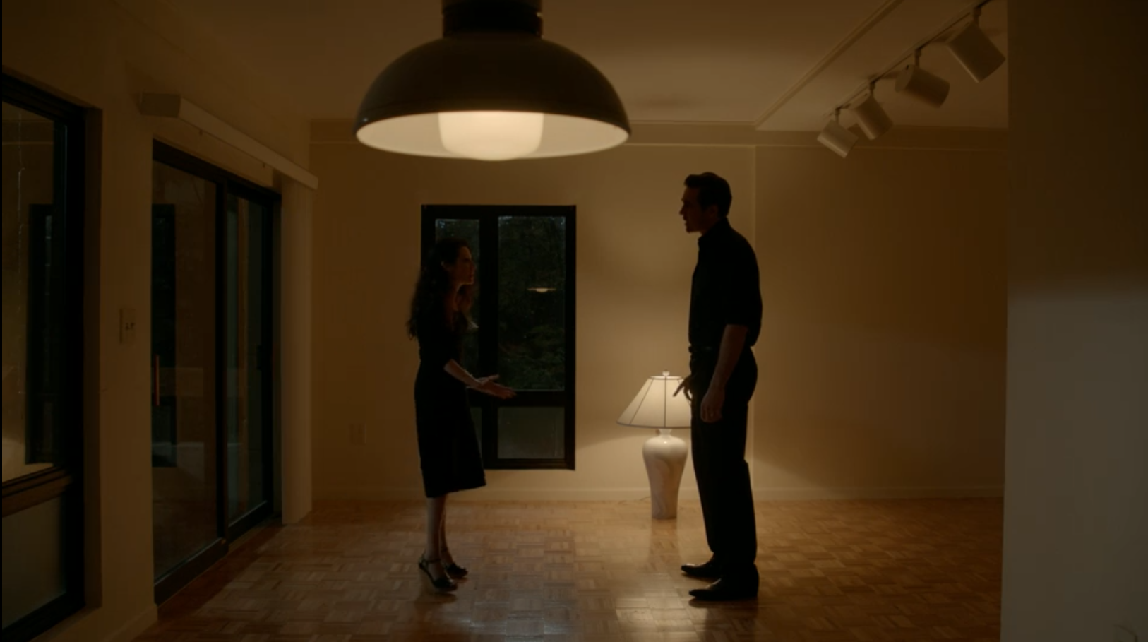 A woman stands in an empty apartment facing a much taller man as they begin to have a verbal fight.