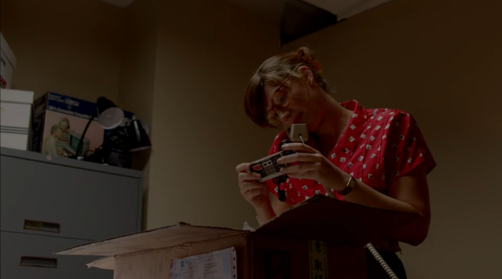 A woman in red top with white dots on it pulls a Nintendo Entertainment System controller out of a cardboard box as she talks on the phone.