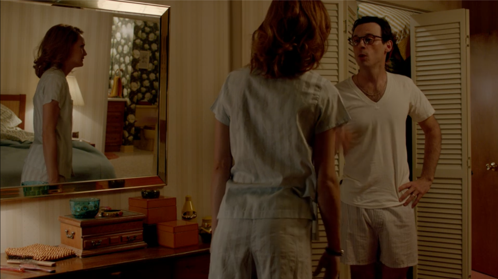 A man in pajamas has an argument with a woman in pajamas. The woman's reflection can be seen in a mirror, so we can see both faces of the people even as the shot is from behind the woman.