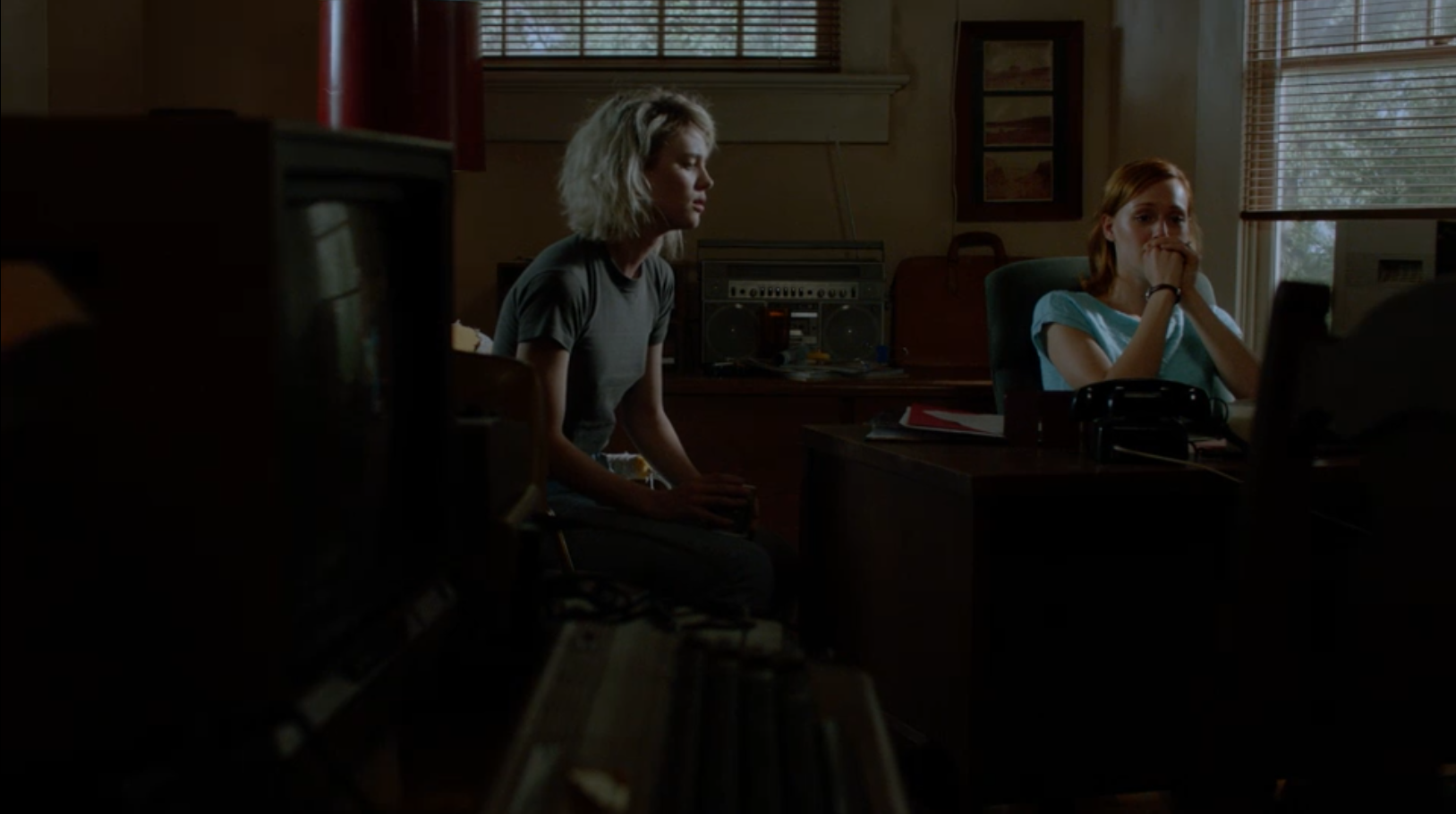 A woman with dyed blond hair sits and talks with a crying woman with red hair in a dark office as it rains outside.