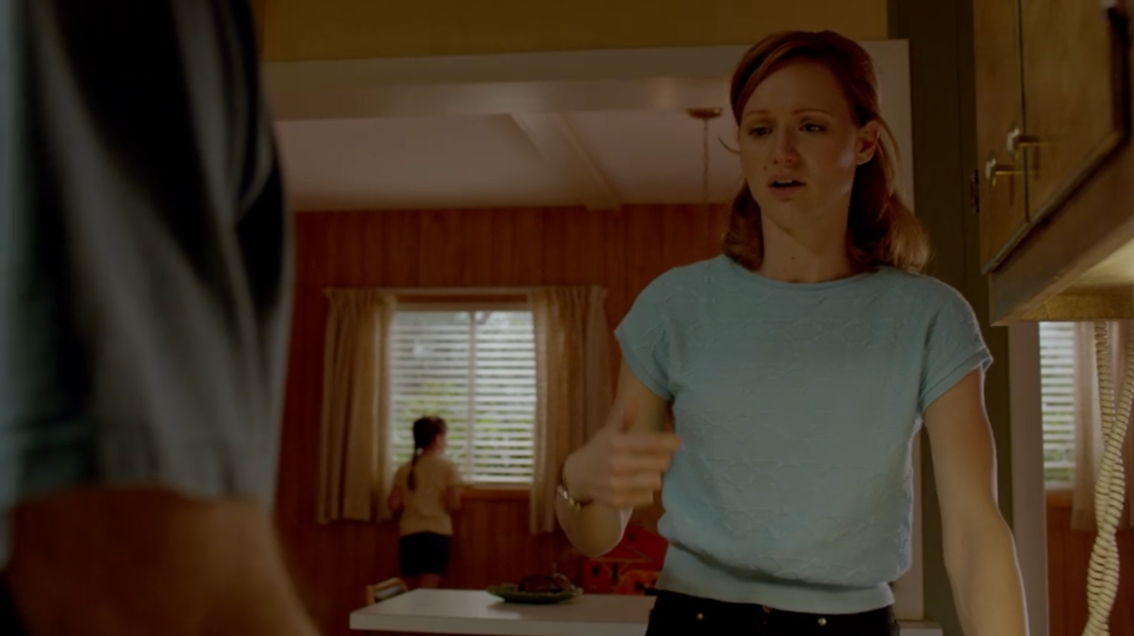 A young girl in the background looks out a window and stares at something, as her parents talk in the foreground.