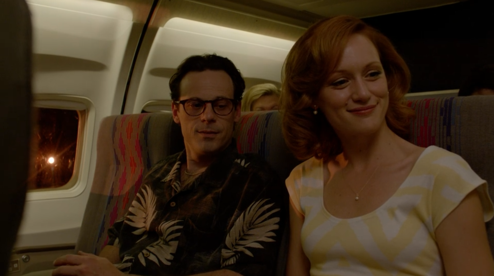 A man in a Hawaiian shirt sits on a plane next to a woman with a yellow and white top. Out the window, there's a bright light.