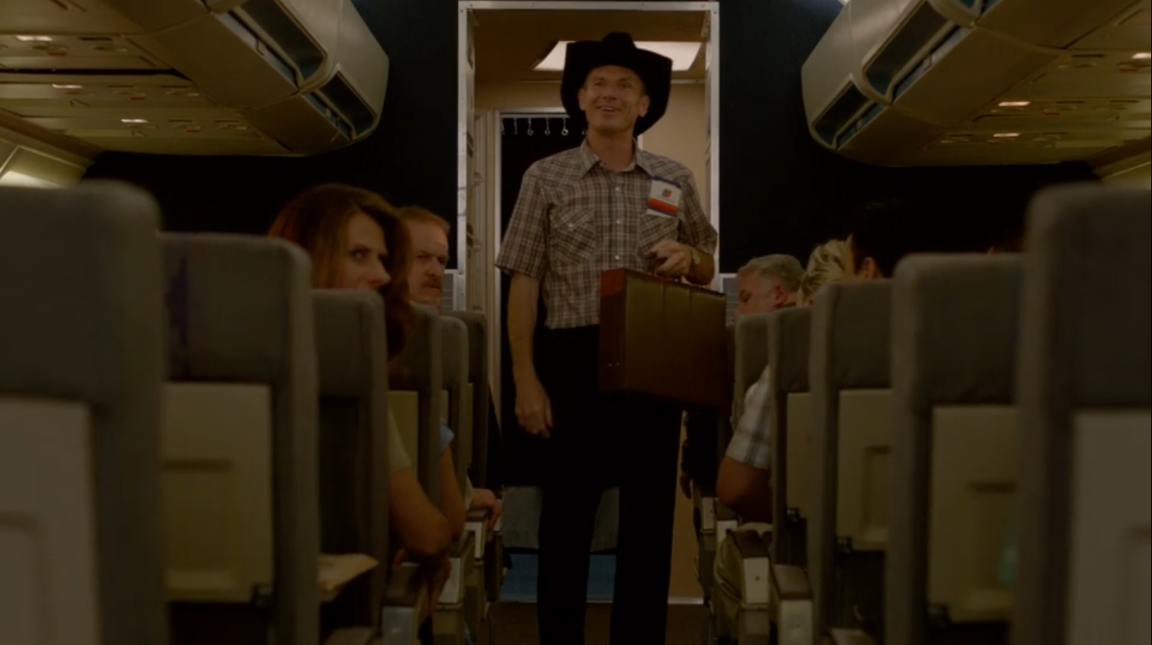 A middle-aged man stands in the aisle of a jetliner wearing a cowboy hat and looking at people cheering him behind the camera.