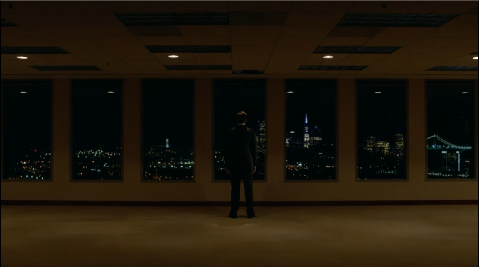 A man stands alone in front of a wall of windows in an empty office as he stares at the San Francisco skyline.