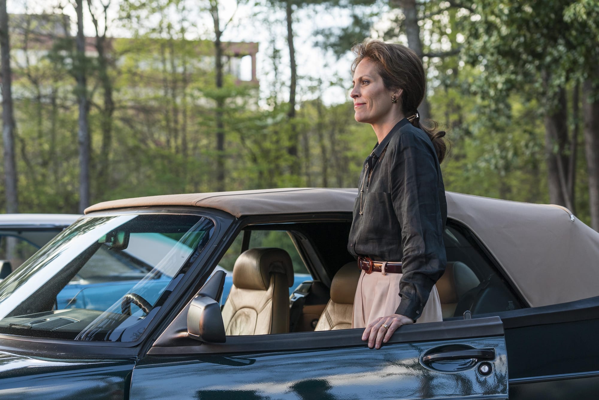 A woman in a gray-brown long sleeve shirt and khaki pants stands next to a car, enjoying the company of someone off screen.