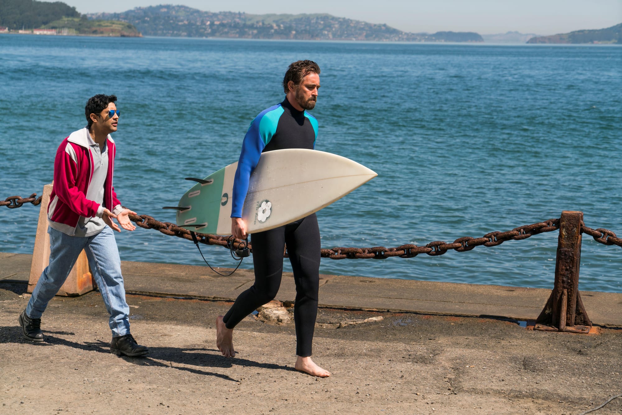 A man in jeans, a red jacket, and sunglasses chases after a barefoot, bearded man in a wetsuit carrying a surfboard. San Francisco Bay is behind them.