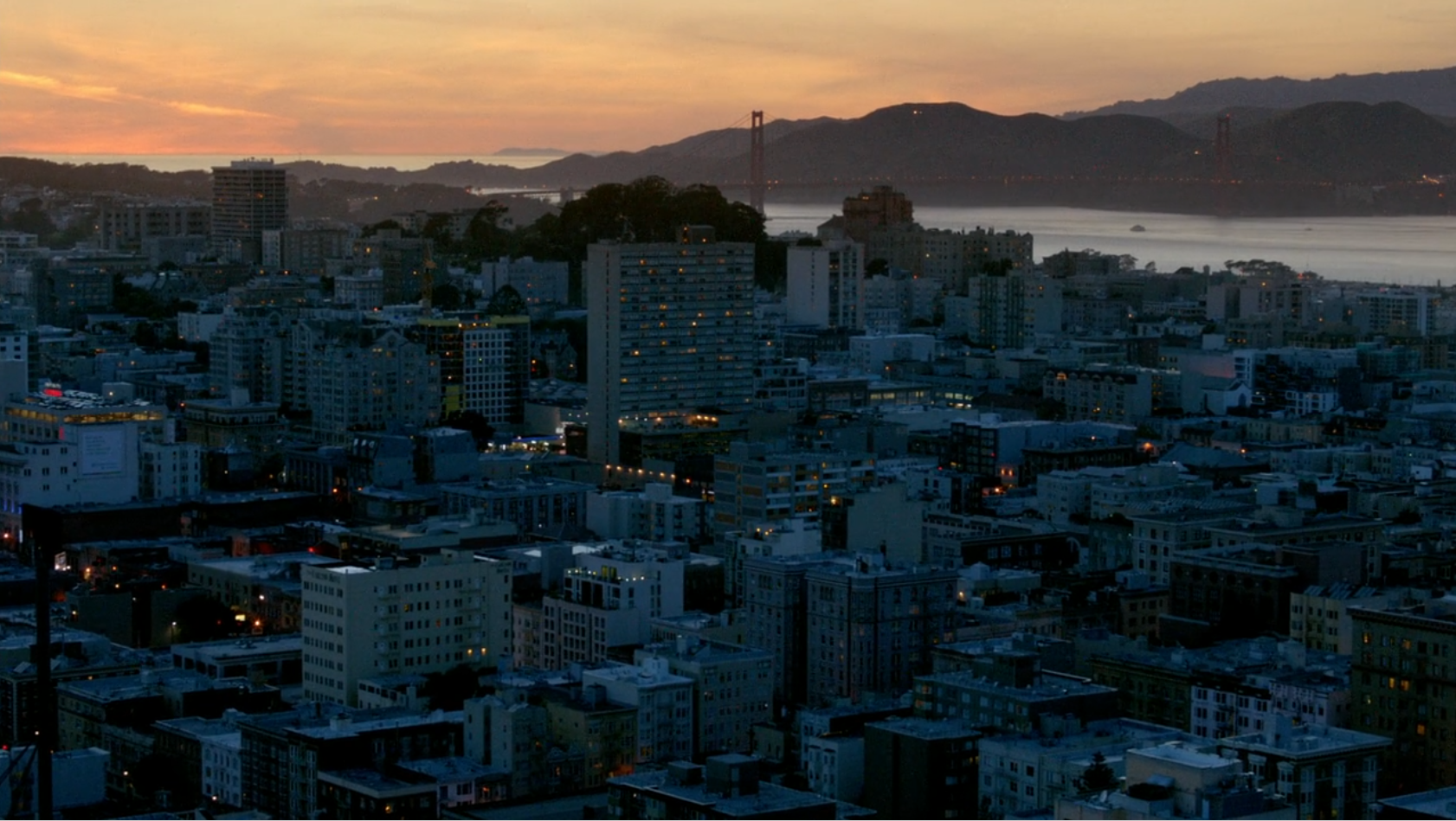 A shot of buildings in San Francisco. The sun is setting, and lights are starting to come on. The Presidio and the Golden Gate bridge are in the background.