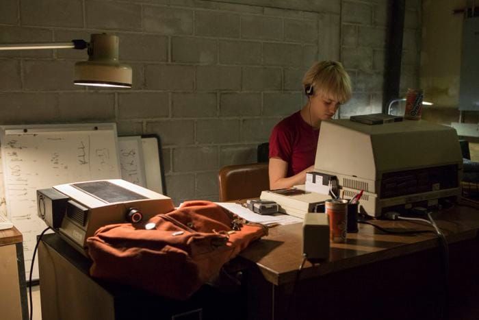 A woman in a red t-shirt sits at a computer in a dimly lit room hard at work on something. She is wearing headphones.