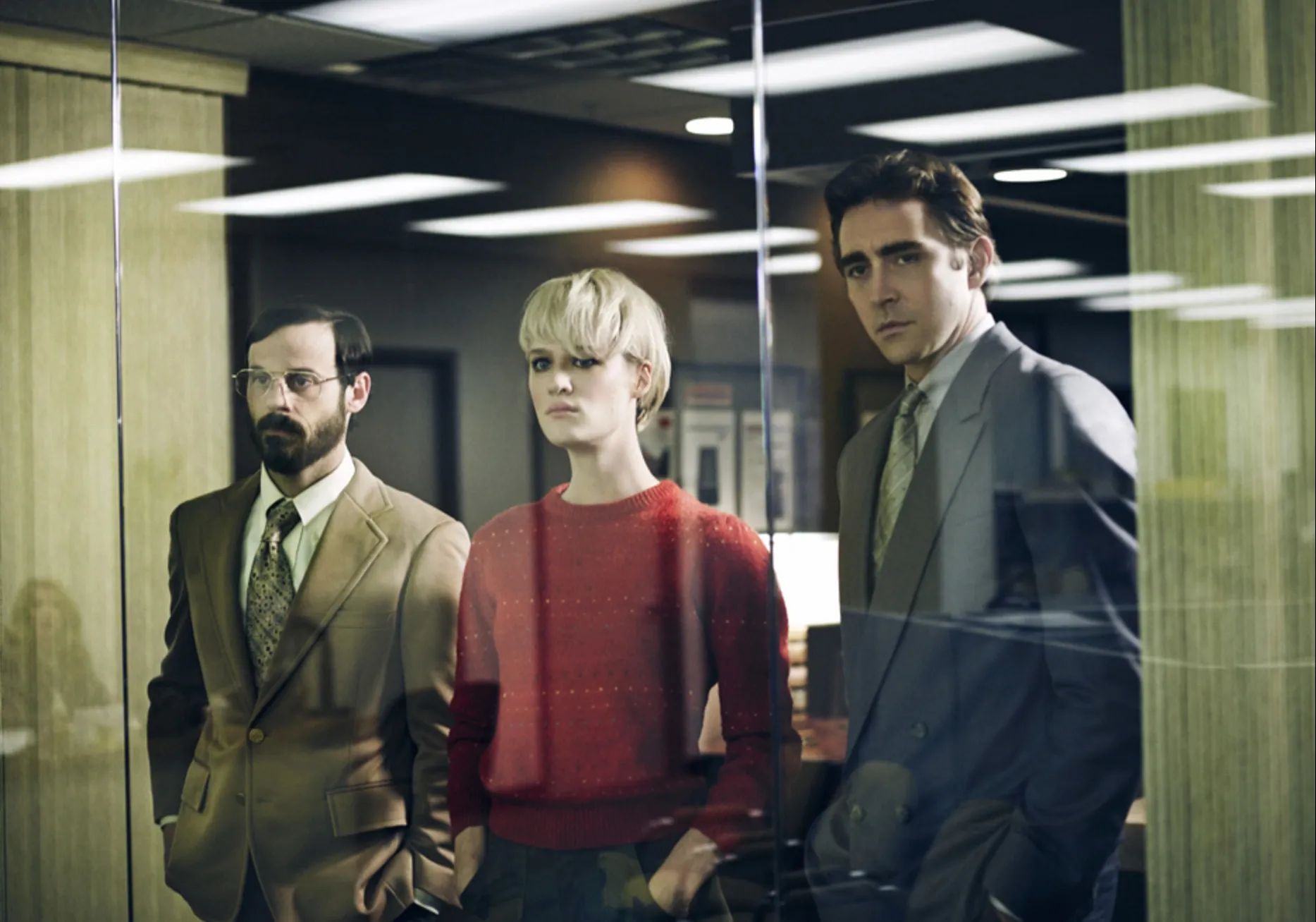 A woman in a red shirt stands between two men in suits in a conference room, looking out at something in the main office.