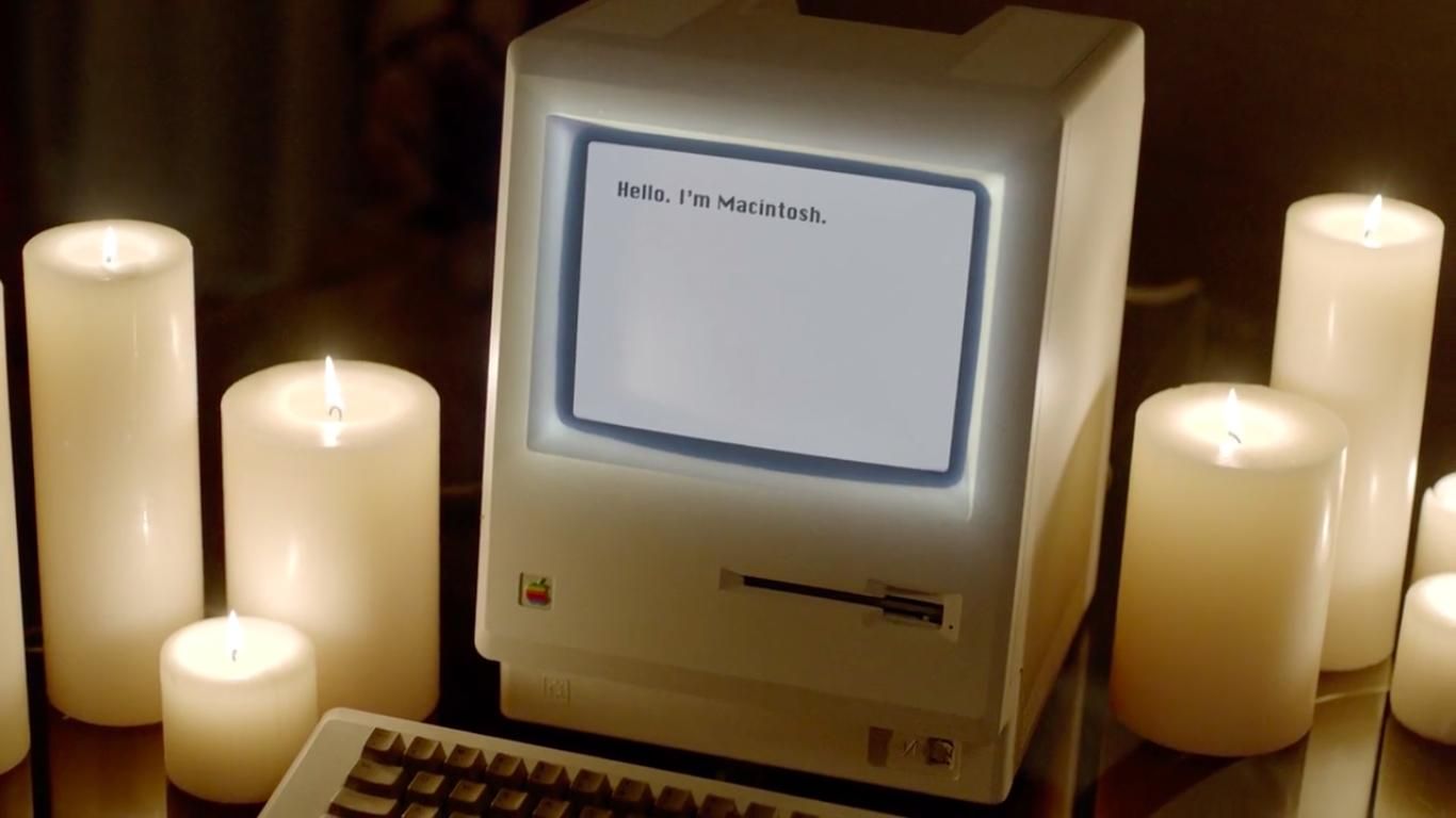 A white vintage Macintosh computer sits in the middle of several lit candles.