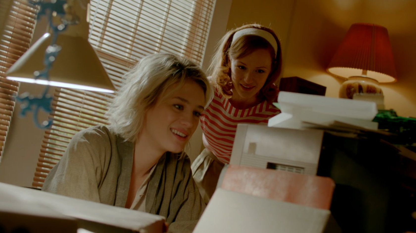 A woman with dyed blonde hair sits a computer as a woman with red hair stands next to her. They are happy at what they see on the screen.