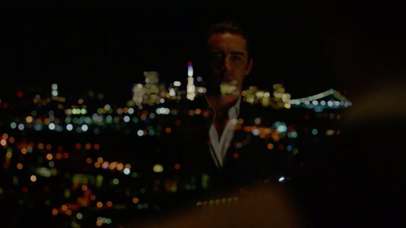 A man's reflection can be seen in a window. He is looking out over the city of San Francisco, which is blurry and lit up in the background.